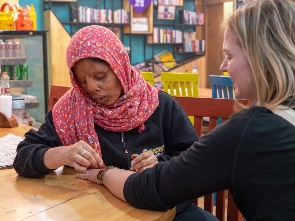 Local woman with visible scarring from an acid attack, applying henna to a female traveller's hand at a table at Sheroes Hangout Cafe in Agra