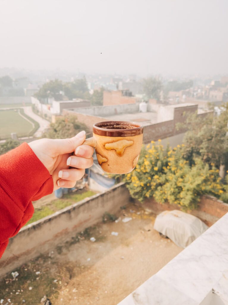 Hand holding a cup of chai on a rooftop terrace overlooking a misty village landscape, capturing a slow morning at a homestay