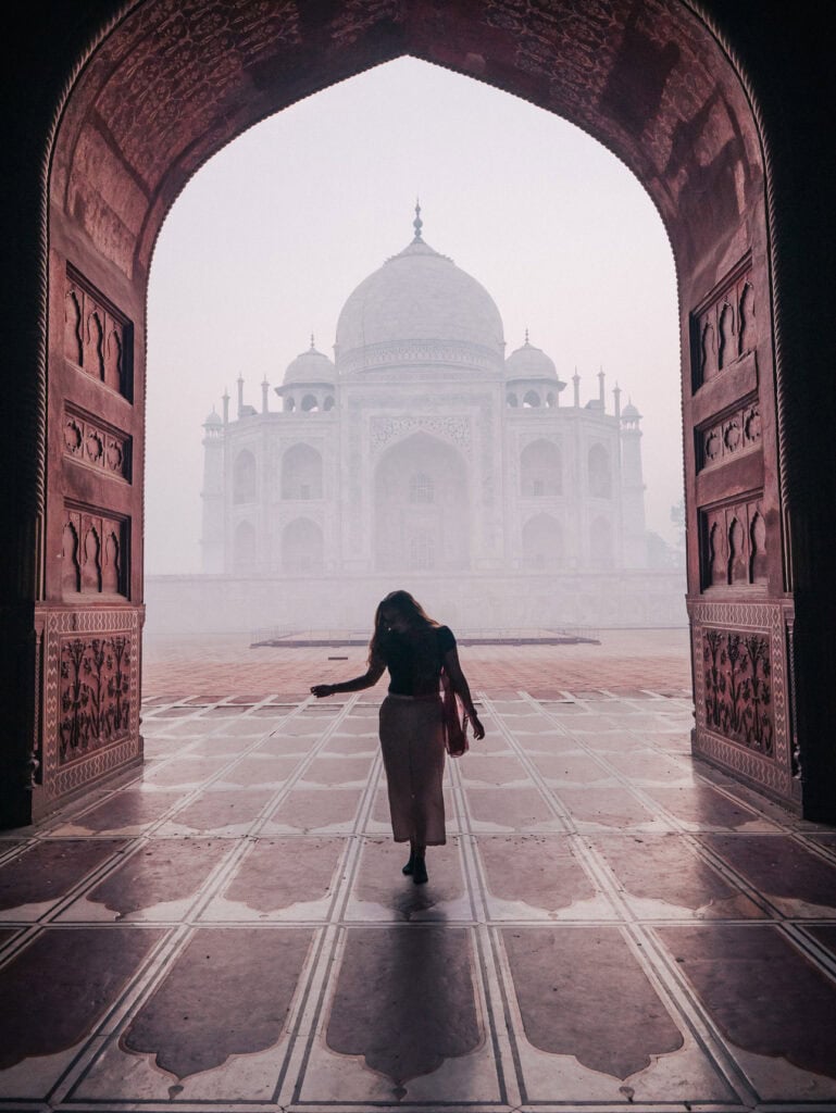 Silhouette of Alexx walking towards the Taj Mahal through a red sandstone gateway in heavy fog