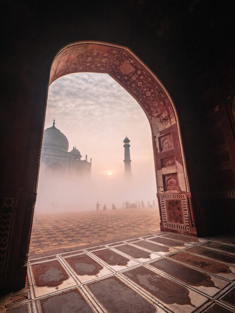 Taj Mahal framed through a large archway in the early morning fog with a few people visible in the distance