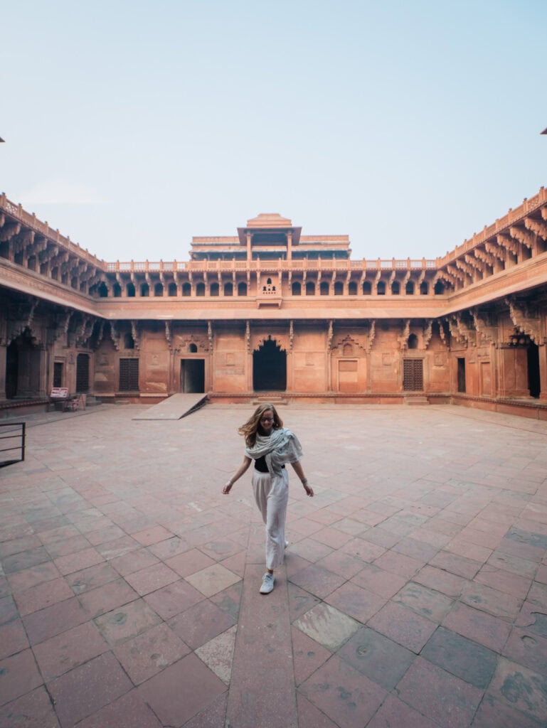 Alexx walking through a large sandstone courtyard at Agra Fort with arched corridors and historic architecture surrounding her