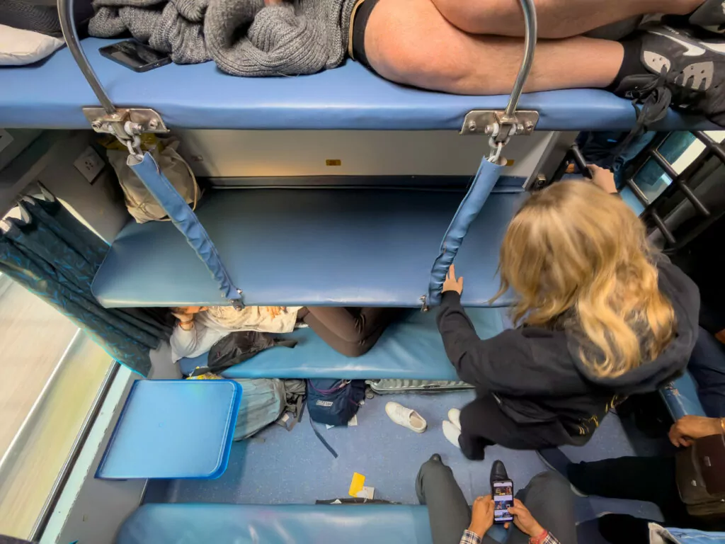 Interior of an AC 3 tier train carriage in India with stacked blue berths and travellers settling in for an overnight journey