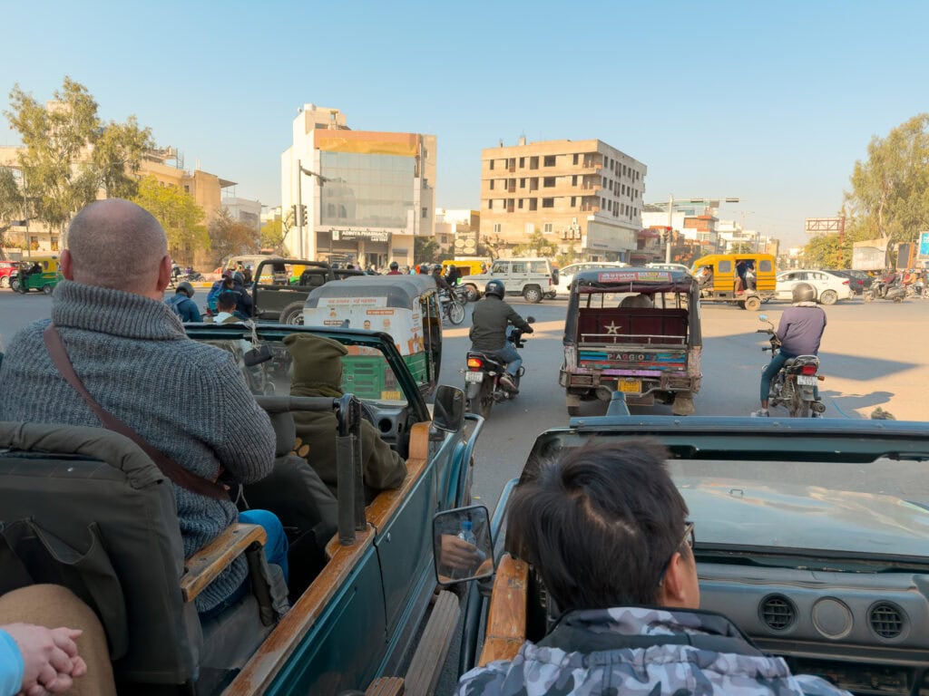 View from the back of an open top jeep driving through traffic in Jaipur with cars, tuk tuks, and buildings ahead