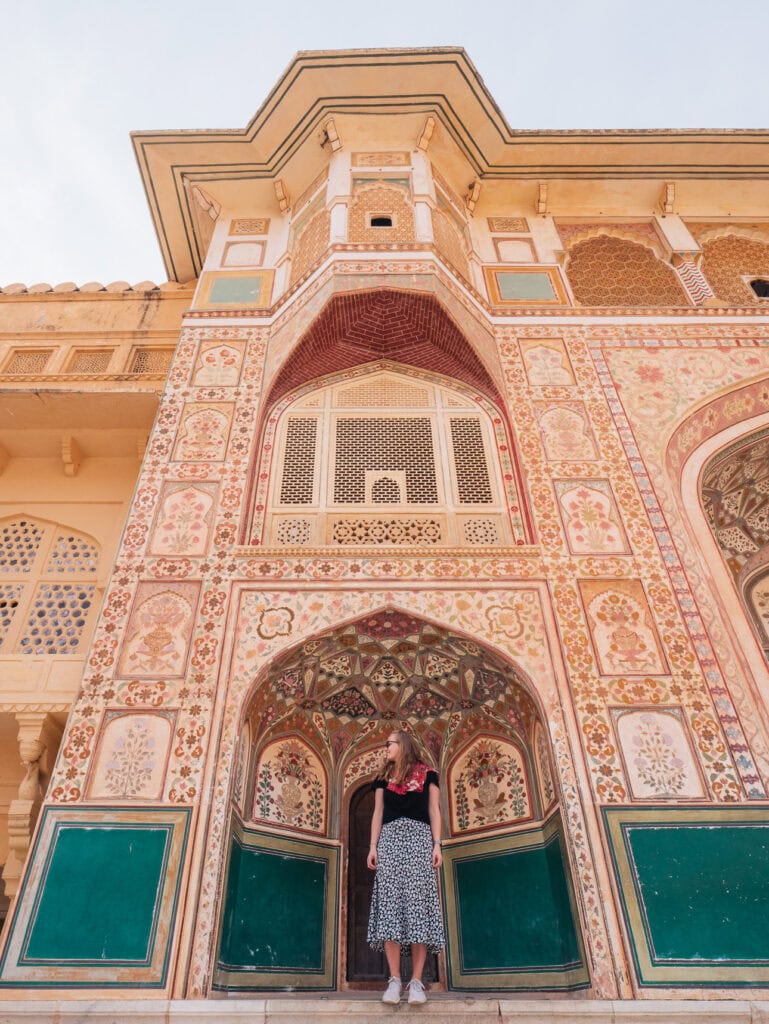 Traveller standing beneath a tall painted palace facade with intricate patterns and a large arched entrance in Jaipur