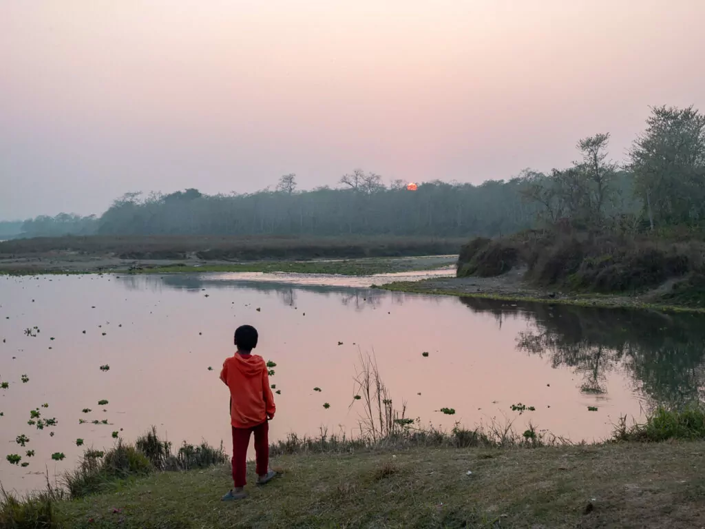 Child standing at the edge of Phewa Lake at dusk with mountains and boats in the background