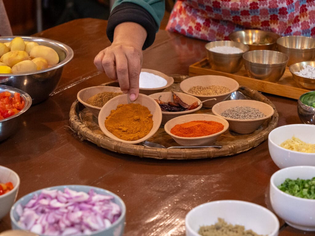 Hands reaching for small bowls of spices and ingredients during a cooking class at Sungabha Seven Sisters in Kathmandu