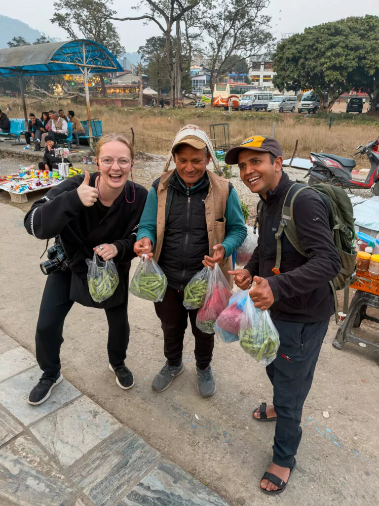 Two travellers and a local vendor giving thumbs up after buying bags of peas on a street in Pokhara