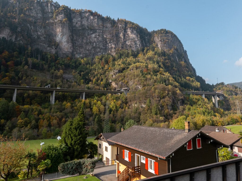A traditional Swiss wooden house tucked beneath a towering cliff face along the Gotthard Railway