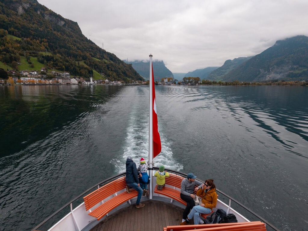 Looking out over the back of a Lake Lucerne boat towards a lakeside Swiss village, with a Swiss flag waving in the breeze