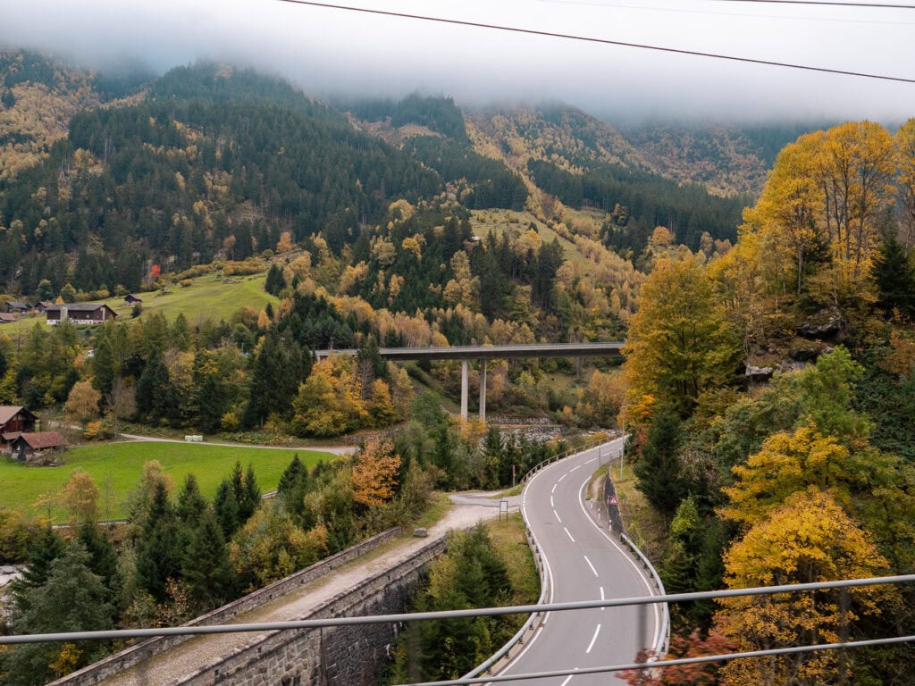 A winding Swiss mountain road passing through a valley of autumn trees, viewed from the Gotthard Panorama Express