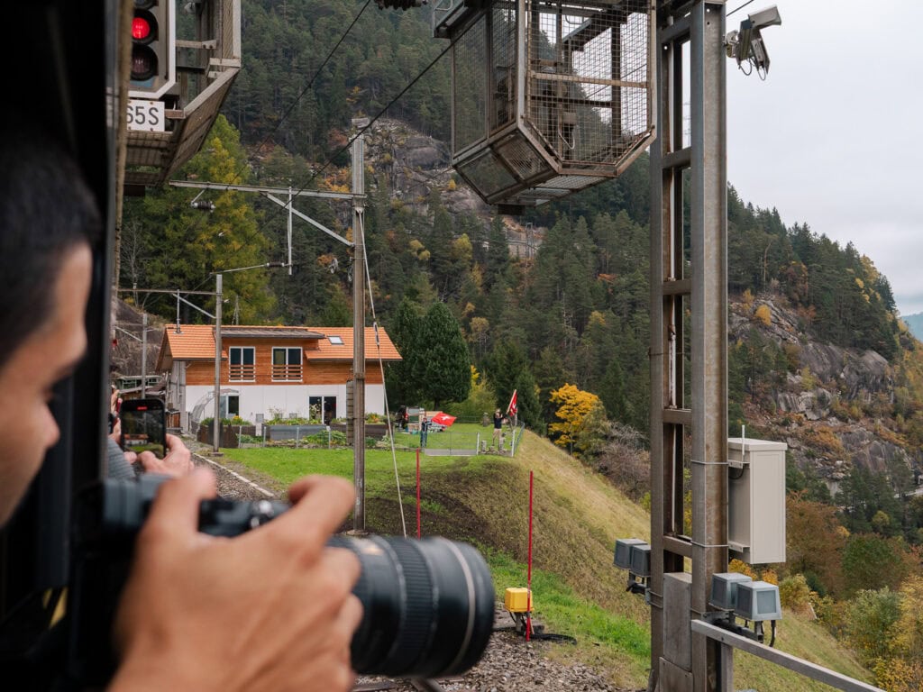 A photographer's hands holding a camera out of the photo coach window of the Gotthard Panorama Express to capture the famous Swiss flag wavers near Wassen