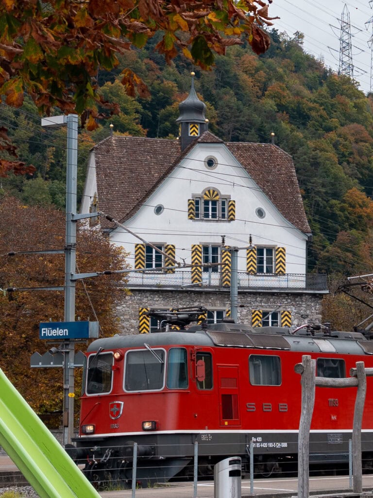 The historic Flüelen station building with its distinctive white facade and green shutters, next to the red driver's cab of the Gotthard Panorama Express train