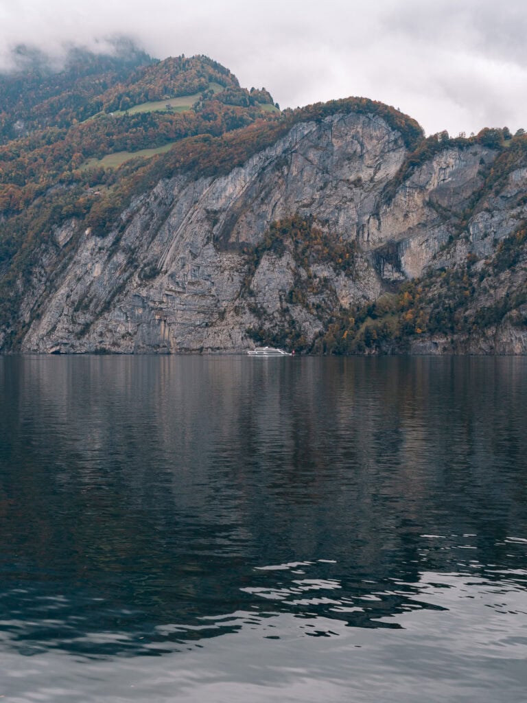 A sheer cliff face meeting the still waters of Lake Lucerne on a cloudy day, with a boat floating in the background