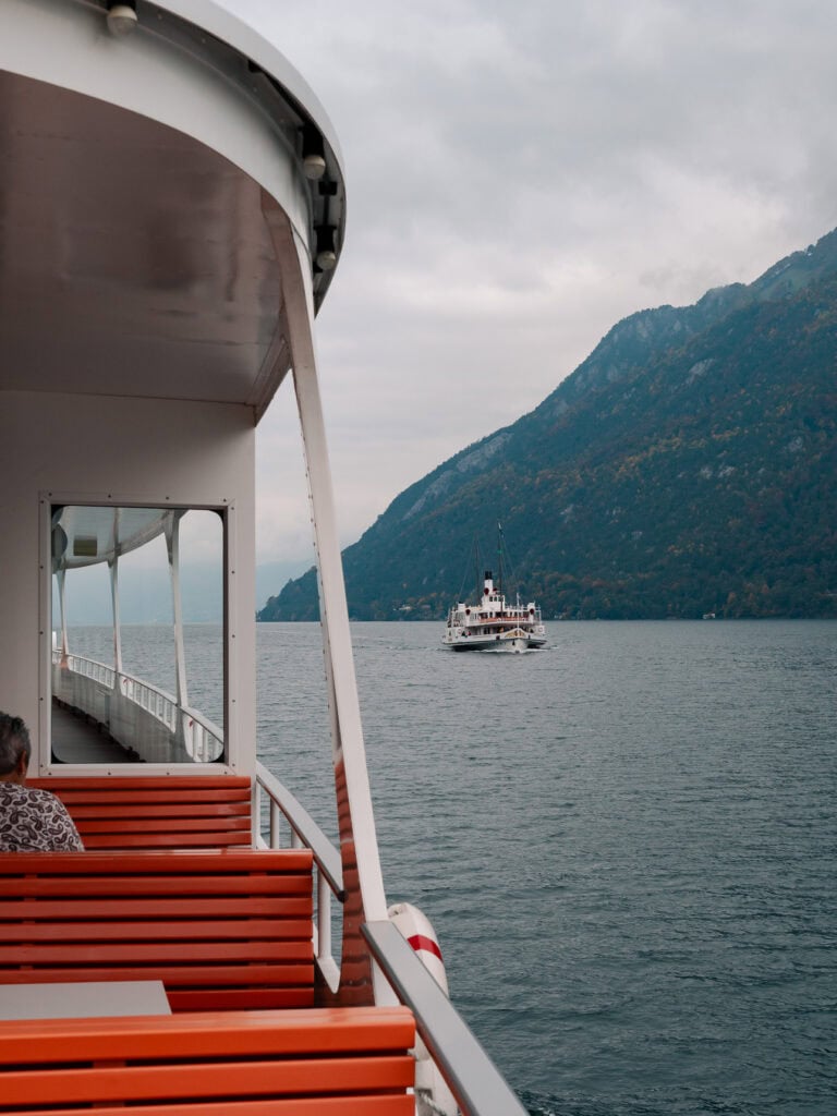 Looking over the side of a Lake Lucerne boat towards a distant vessel and misty mountains on an overcast day