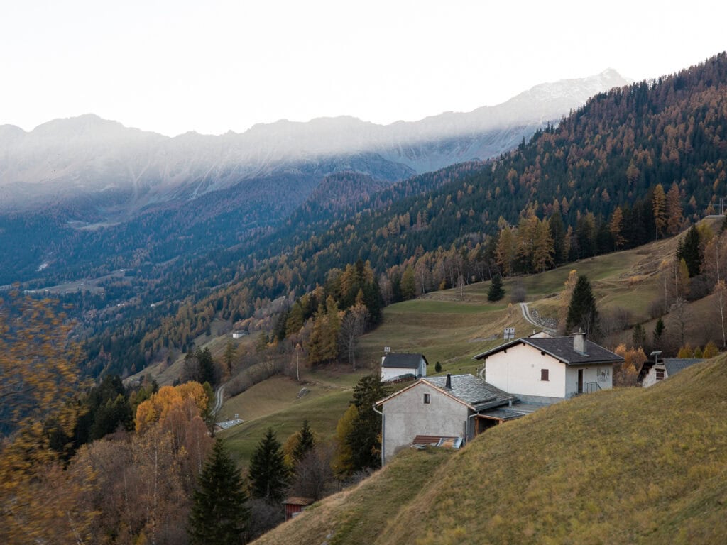 View from the Bernina Express train window at dusk, looking out over an alpine valley with rustic white farmhouses on a hillside, autumn-coloured forests and snow-dusted mountain peaks partially obscured by low cloud