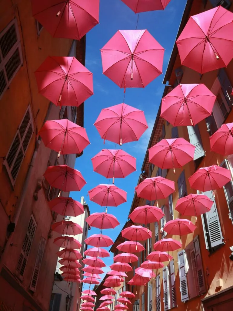 Pink umbrellas hang above a narrow street between orange buildings. The bright installation adds colour to a Riviera style village walk.