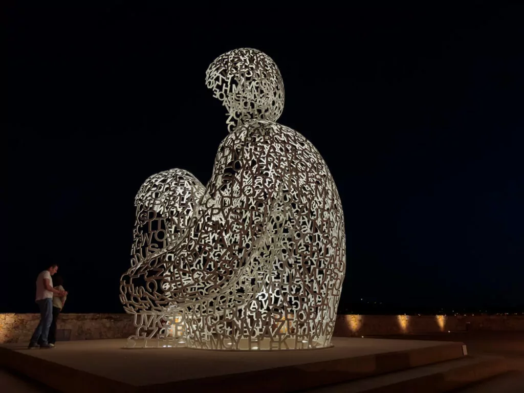 The illuminated Le Nomade sculpture in Antibes glows against a dark night sky. The seated figure is made from white metal letters.
