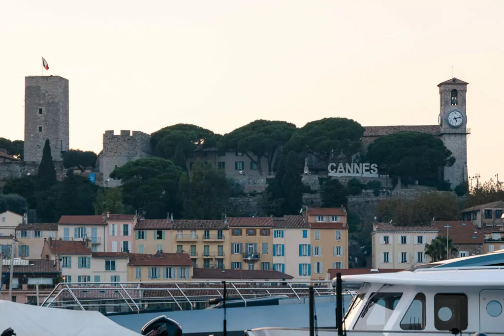 Cannes old town rises above the harbour with a clock tower and hillside sign. The sign reads "Cannes".