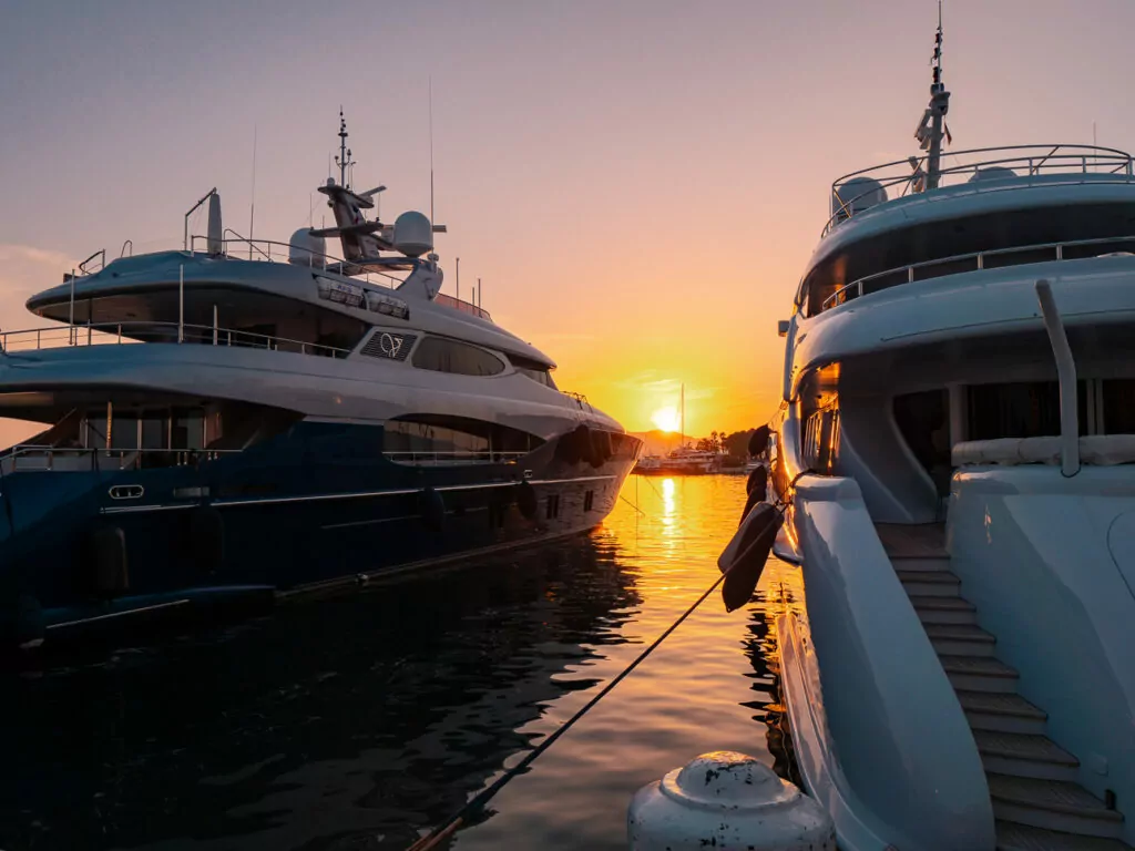 Superyachts sit in Cannes harbour at sunset with orange light reflecting between the boats. The marina shows the glitzy side of the Riviera coast.