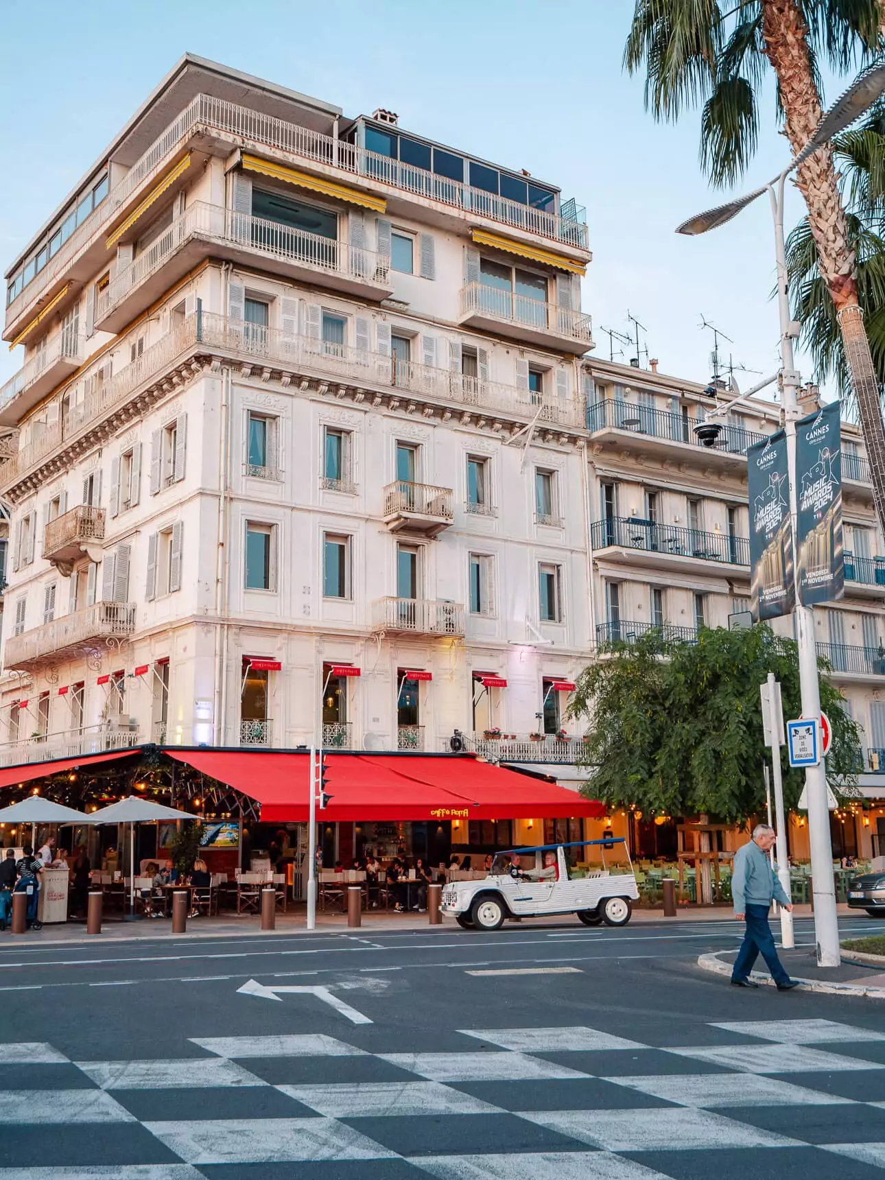 A Cannes street corner with a red awning cafe. Palm trees. Hotel balconies. And banners for the city beside the road