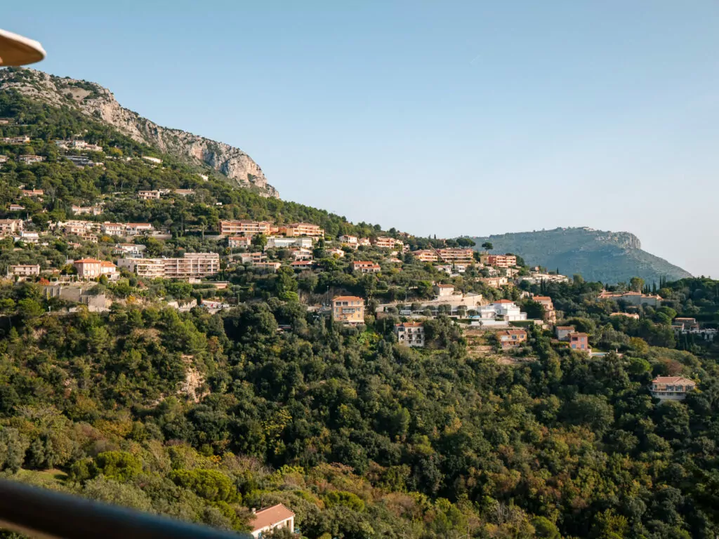 Hillside homes near Èze climb through dense green trees toward rocky cliffs. The steep terrain makes this a scenic but slightly more complex car free stop.