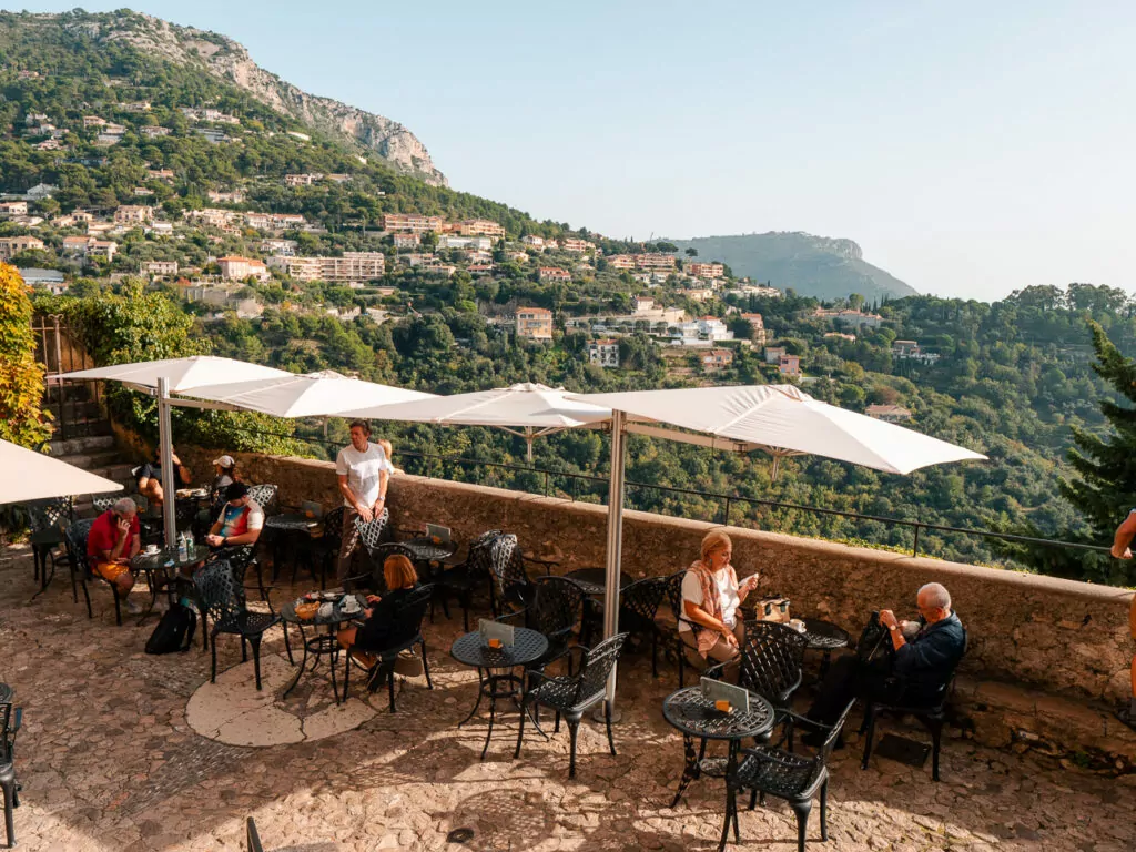 A cafe terrace in Èze overlooks green hills and the Mediterranean coast. This is a scenic place to pause during a public transport day trip.