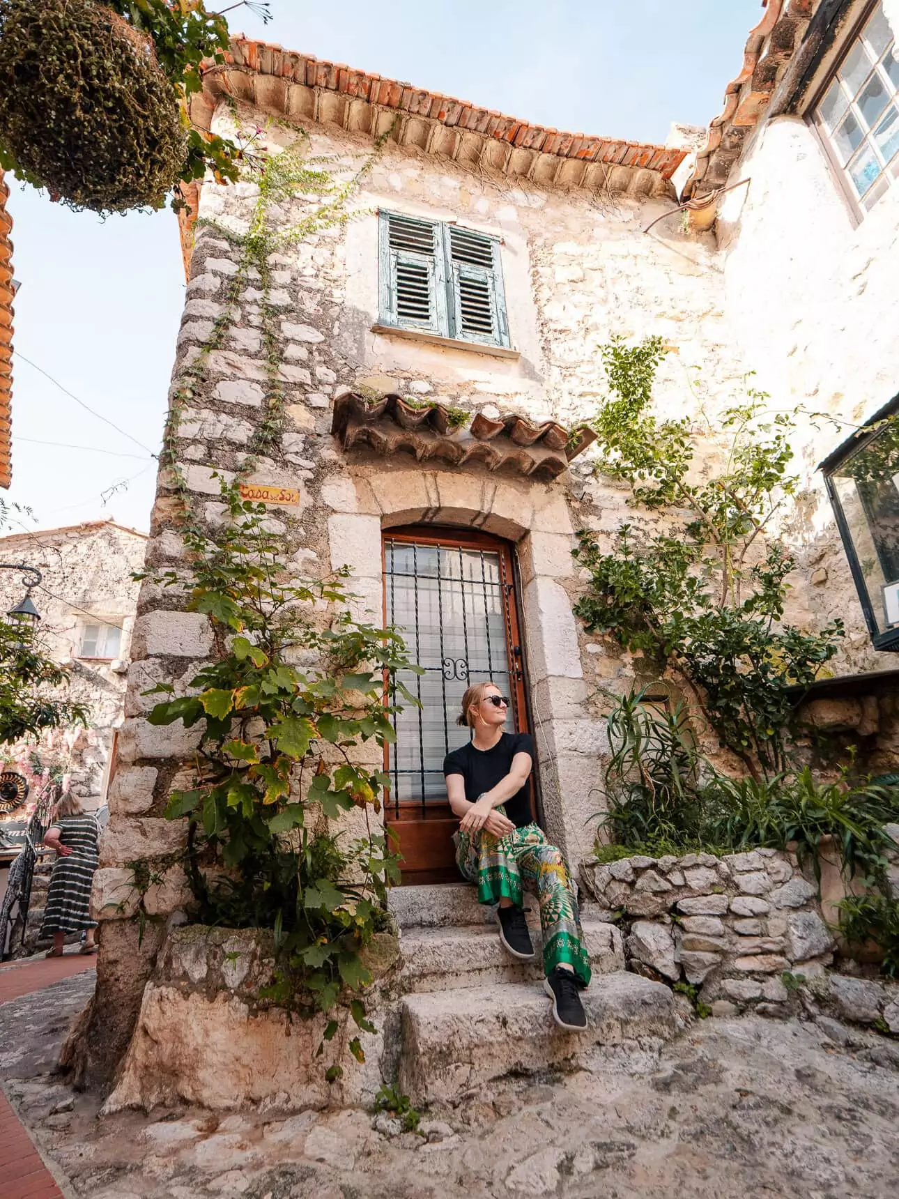 A woman sits on stone steps beside an old house in Èze village. The cobbled lane and climbing plants show the medieval charm of this hilltop stop.