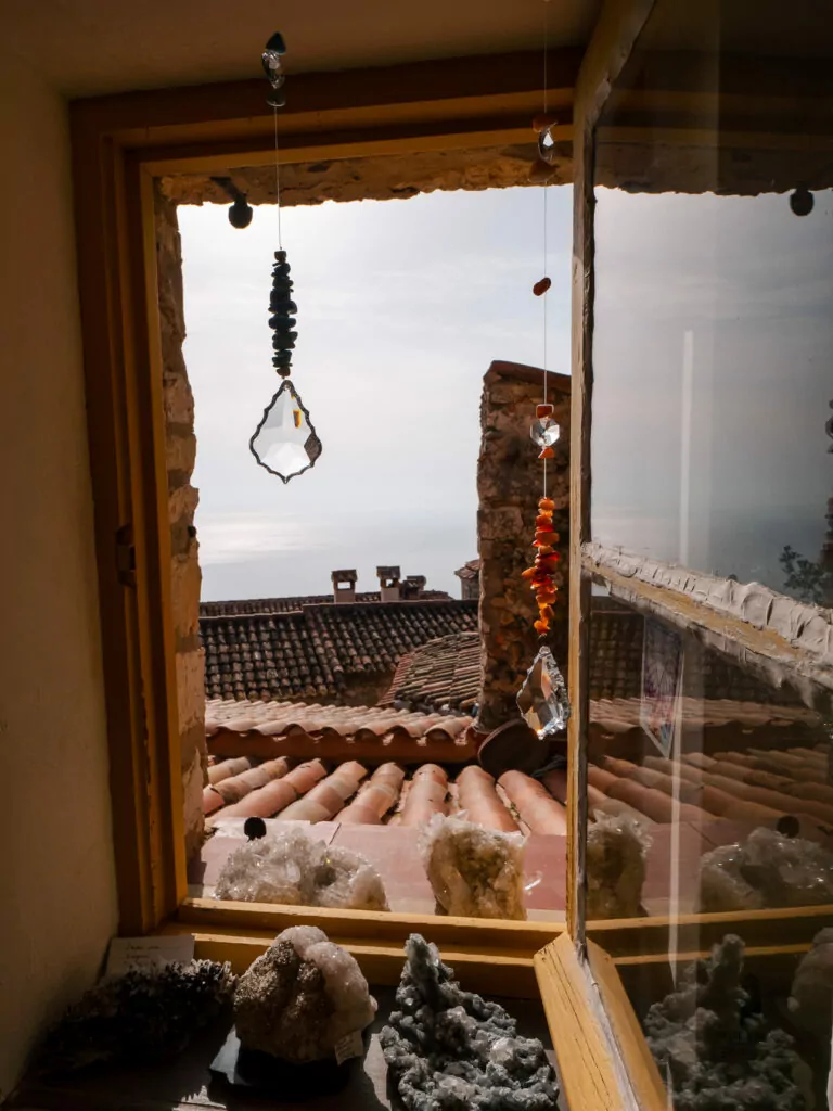 Crystals hang in a shop window in Èze with terracotta rooftops and hazy sea beyond. The small display captures the village craft shops and steep coastal views.