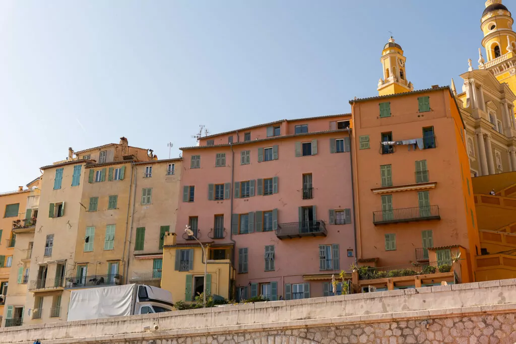 Pastel waterfront buildings in Menton sit below the church tower and bright orange ramparts. The old town rises directly above the sea.
