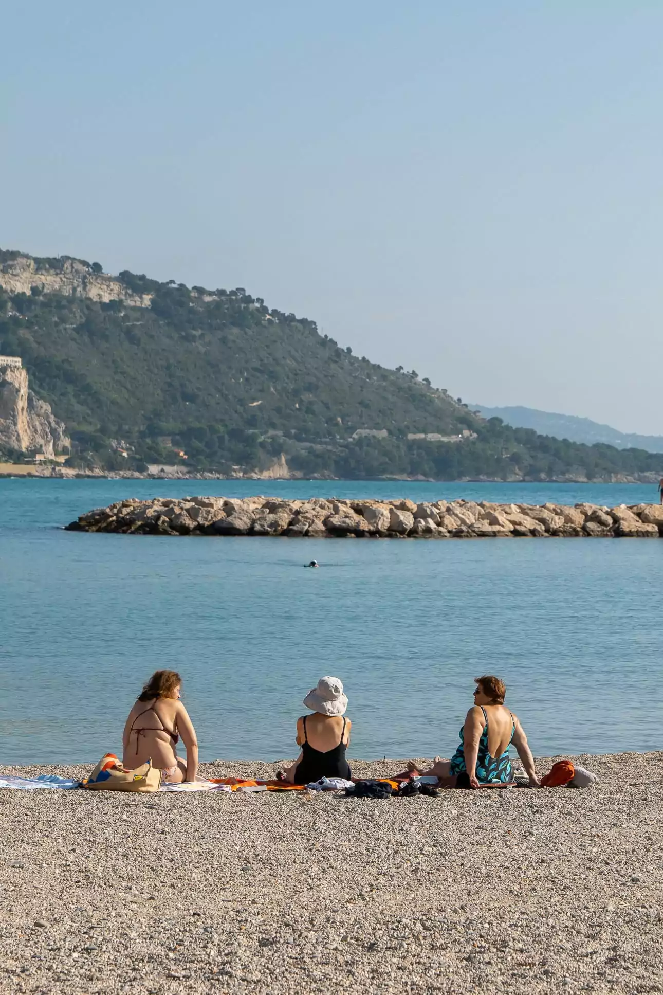 Three people sit on towels on a pebble beach in Menton with cliffs and blue sea behind them