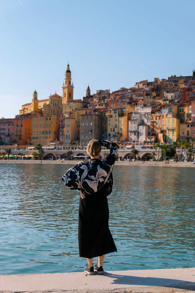 A woman looks across the water toward Menton old town with pastel buildings glowing in the sun. It is one of the most colourful stops on a French Riviera without a car route.