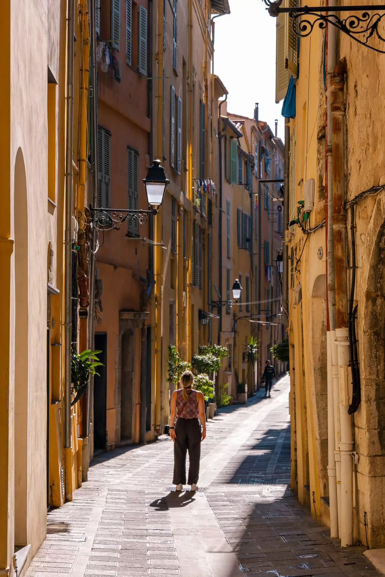 A person walks down a narrow sunlit street in Menton old town. Tall yellow and orange buildings line the lane
