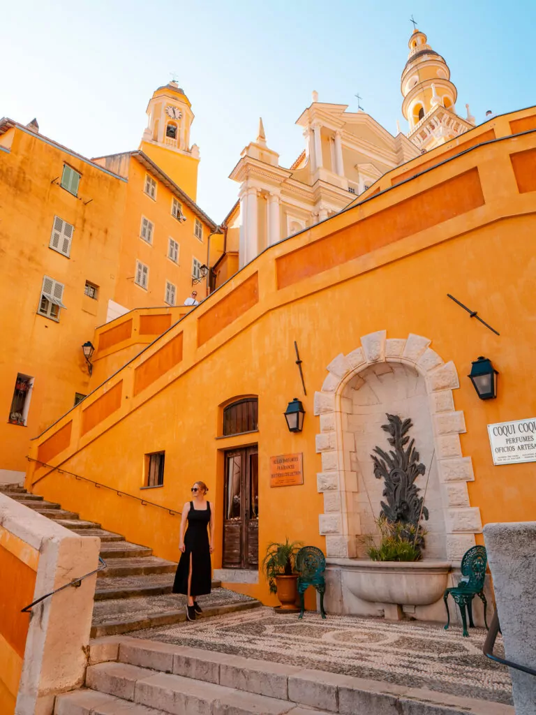 A woman stands on the yellow Rampes Saint Michel below the church towers in Menton. The bright stairs and old town buildings make this a standout photo spot.