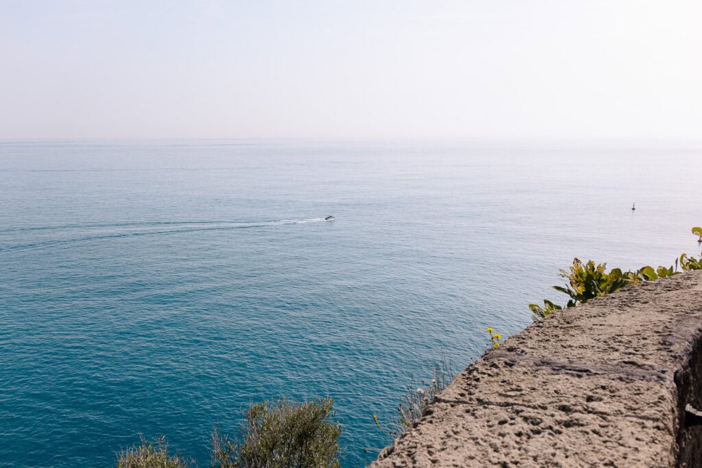 A small boat crosses calm blue water below a stone viewpoint in Monaco. The open sea view adds a scenic break to a car free Riviera day trip