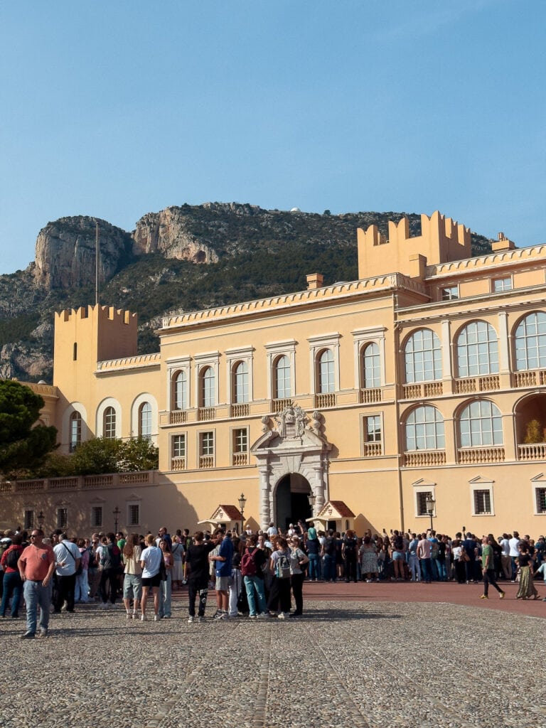 Crowds gather outside the Prince Palace in Monaco with rocky hills behind it. This palace square is easy to visit on a day trip from Nice by train