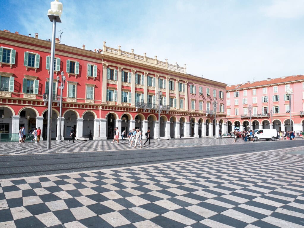Place Masséna in Nice with red arcaded buildings and a black and white patterned square. People cross the wide plaza in the sun