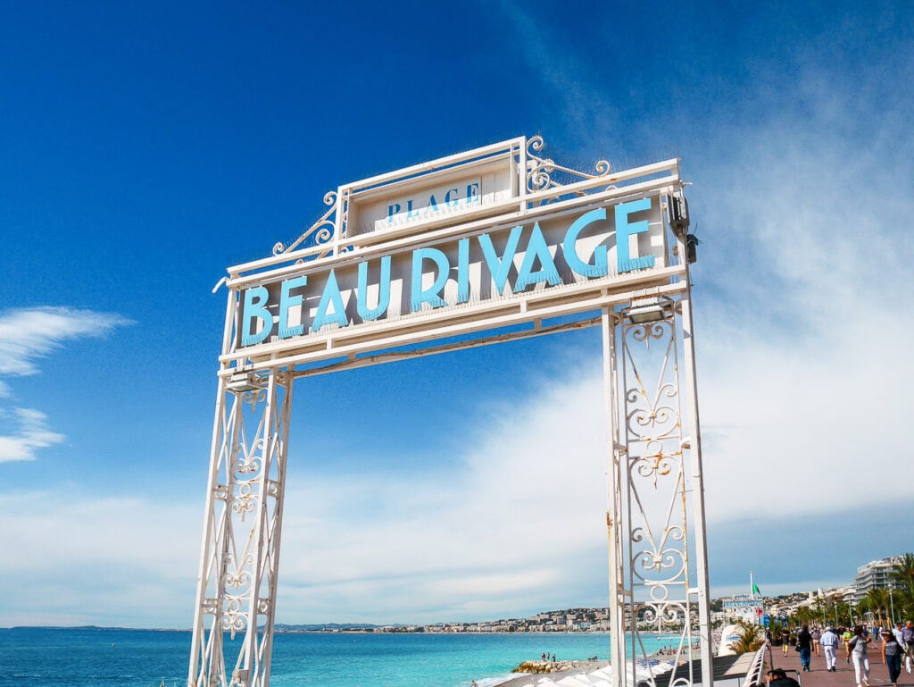 The white and blue beach sign for Beau Rivage stands above turquoise water in Nice. Text reads "Plage Beau Rivage"