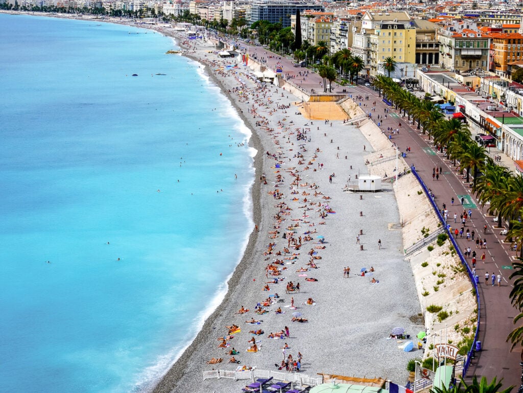 Nice beach seen from above with turquoise water. Pebble shoreline. Promenade des Anglais. And crowds sunbathing along the bay.