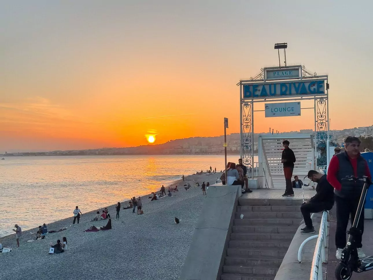 Sunset over Nice beach with people sitting on the pebbles beside the water. The beach entrance sign reads "Plage Beau Rivage Lounge"