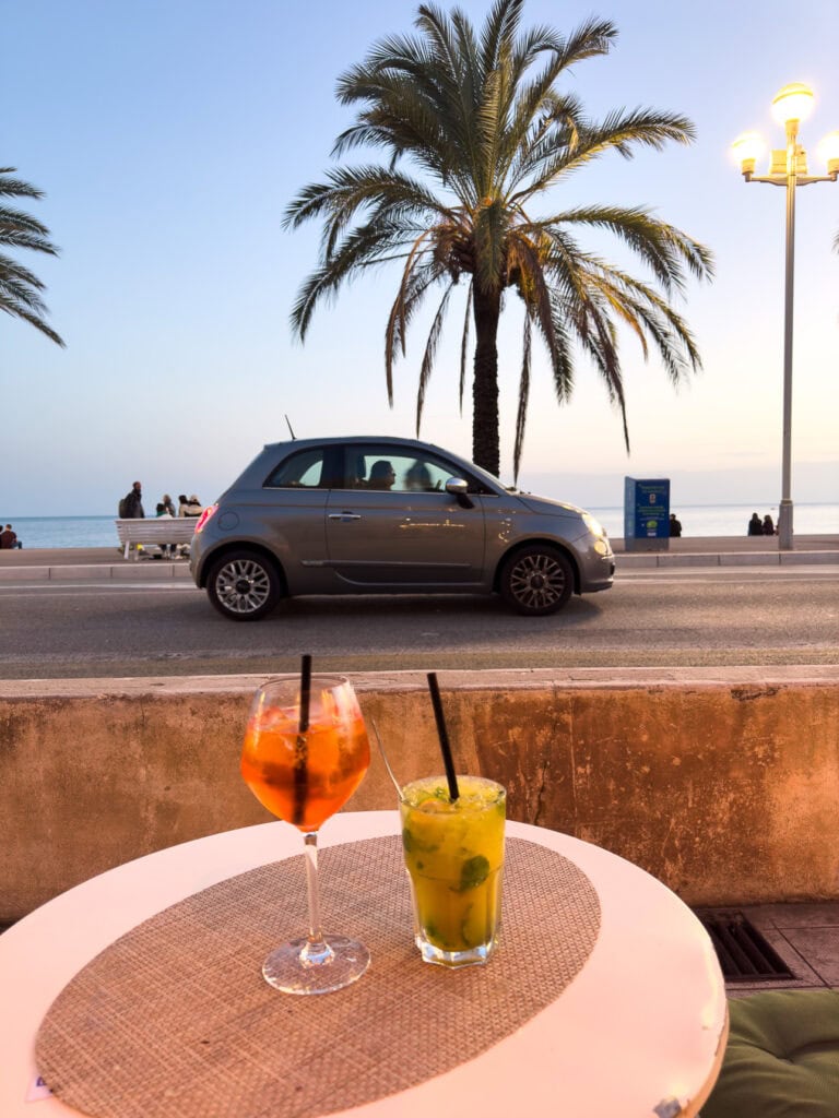 Two drinks sit on an outdoor table beside the Promenade des Anglais in Nice. A small Fiat 500 car and palm tree frame the sunset view over the sea.