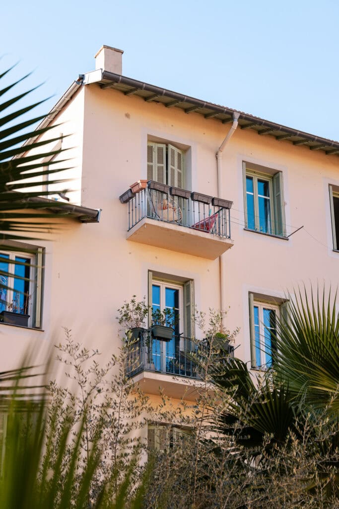 A pale pink building with small balconies sits behind palm leaves in Nice. The soft colours and shutters give the stay a relaxed Riviera feel