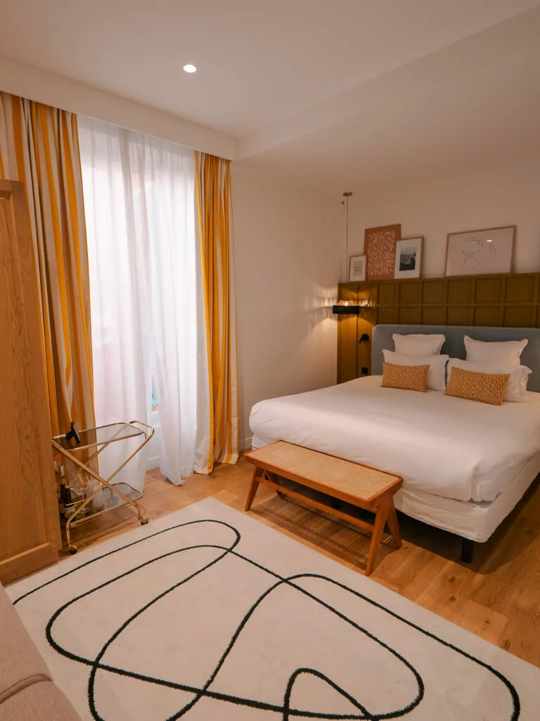 A boutique hotel room at Maison Durante in Nice with a white bed. Mustard curtains. Wood furniture. And a patterned rug