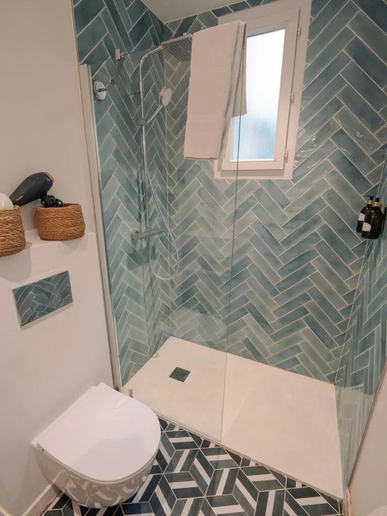 A compact hotel bathroom in Nice with teal herringbone tiles. A glass shower. A towel by the window. And a toilet beside the vanity.