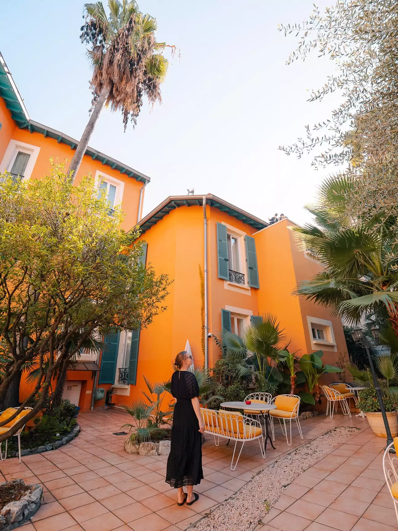 A woman in a black dress stands in a sunny orange hotel courtyard in Nice. The courtyard has palms. Yellow chairs. And blue window shutters