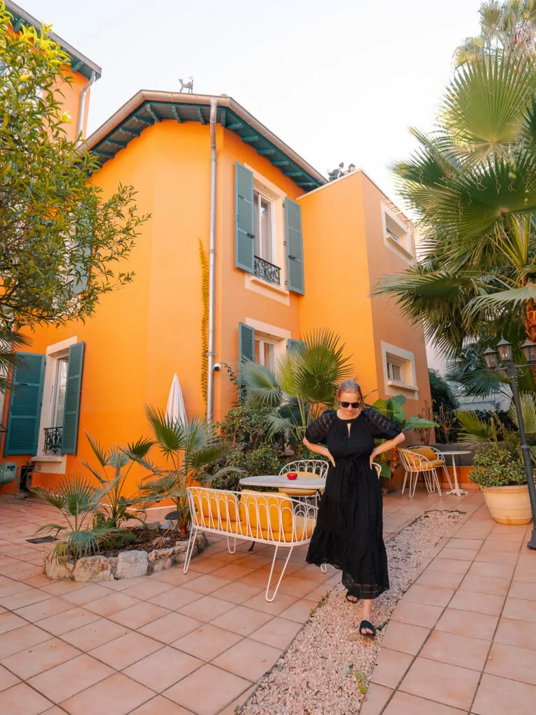 A woman walks through an orange courtyard in Nice surrounded by palms and patio tables at Maison Durante. The setting shows a cheerful hotel option for a car free Riviera base