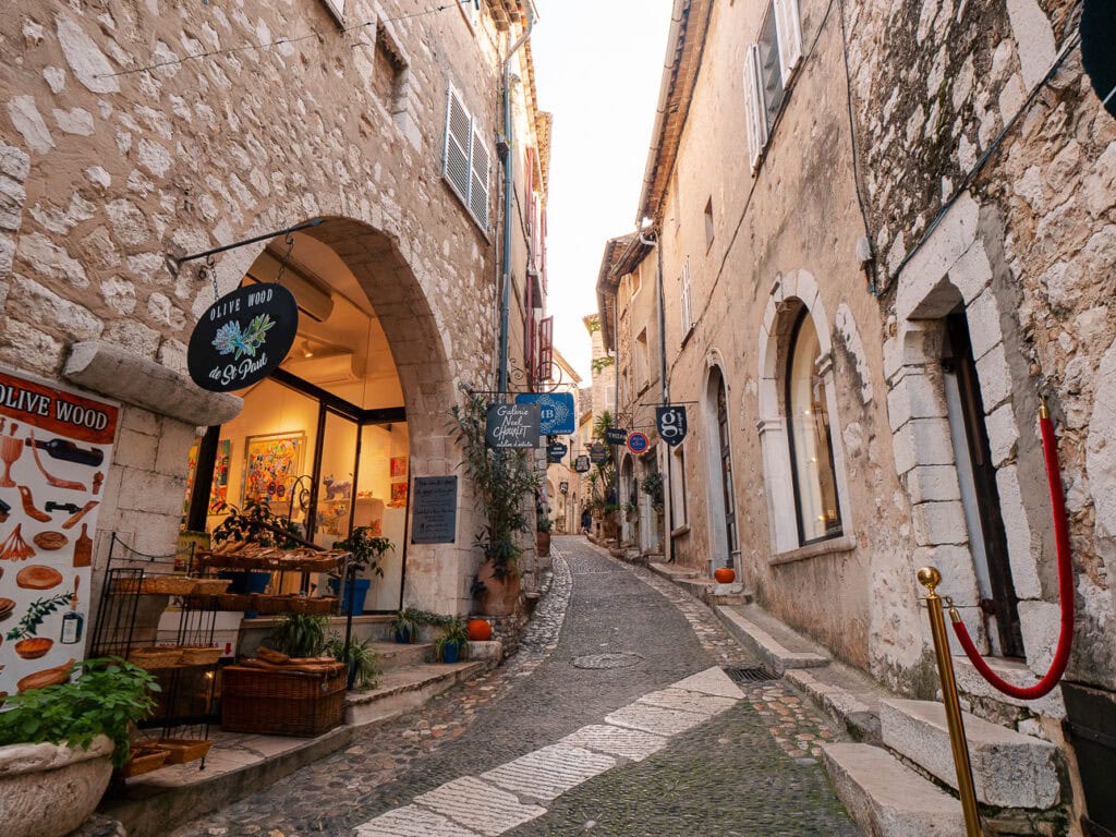 A cobbled lane in Saint Paul de Vence passes stone buildings and small shops. Visible signs read "Olive Wood" and "Galerie"