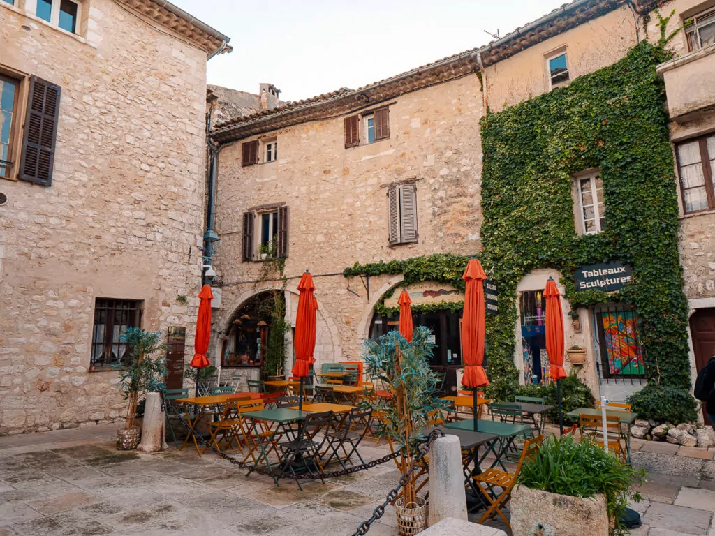 A quiet courtyard in Saint Paul de Vence has orange umbrellas and tables beneath old stone buildings. A sign reads "Tableaux Sculptures".