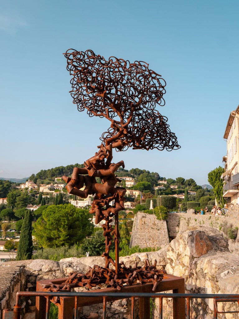 A rust coloured metal sculpture stands on the ramparts in Saint Paul de Vence. The countryside and stone walls behind it show the village art scene.