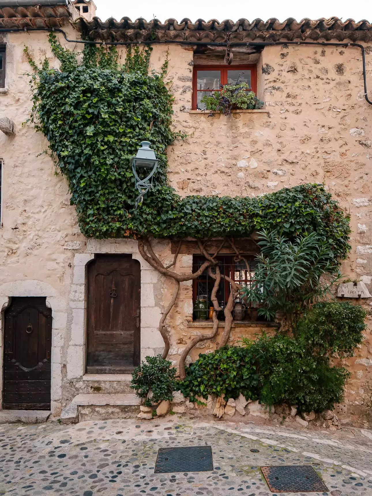 An ivy covered stone house with wooden doors and shuttered windows in Saint Paul de Vence. The old village streets show the inland side of a car free Côte dAzur itinerary