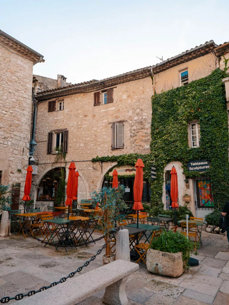 A cafe courtyard in Saint Paul de Vence sits beneath old stone buildings and orange umbrellas. A sign reads "Tableaux Sculptures".